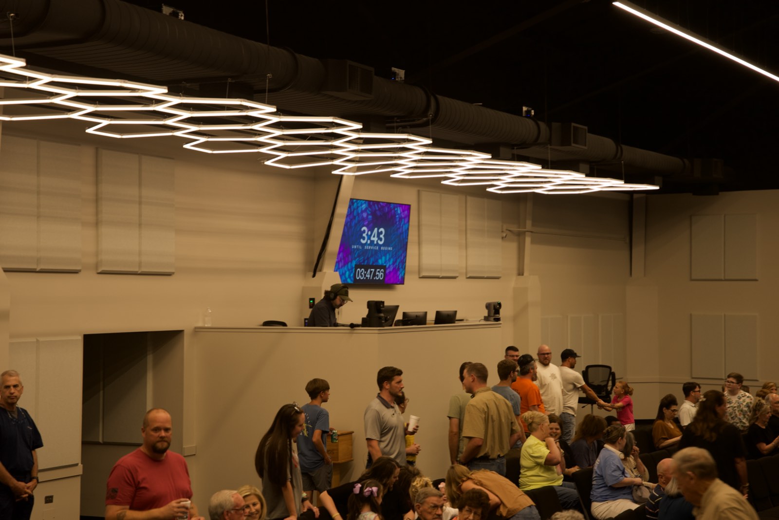 Hexagon light fixture above the tech booth at First Baptist Church Pontotoc, with acoustic panels on the wall and people gathering before service