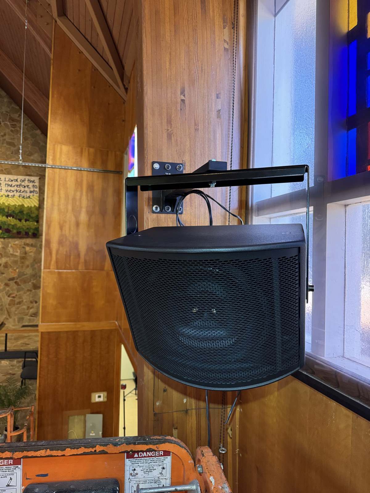Compact black loudspeaker on a wall bracket beside a stained glass window at St. John's Lutheran Church