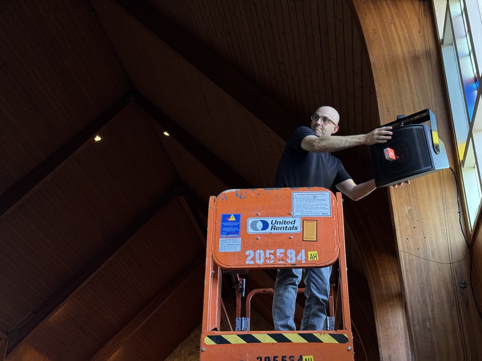 Churchfront installer on a scissor lift mounting a compact speaker high on the wood-paneled wall at St. John's Lutheran Church
